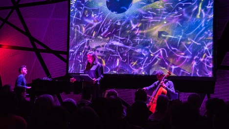 Three musicians performing on stage in front of a screen with artwork on it