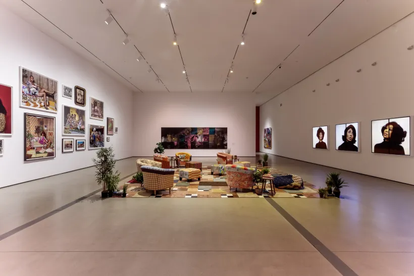 Gallery shot of Mickalene Thomas All About Love Exhibition featuring a living room set up with a photo gallery on the left wall and four video screens on the right wall